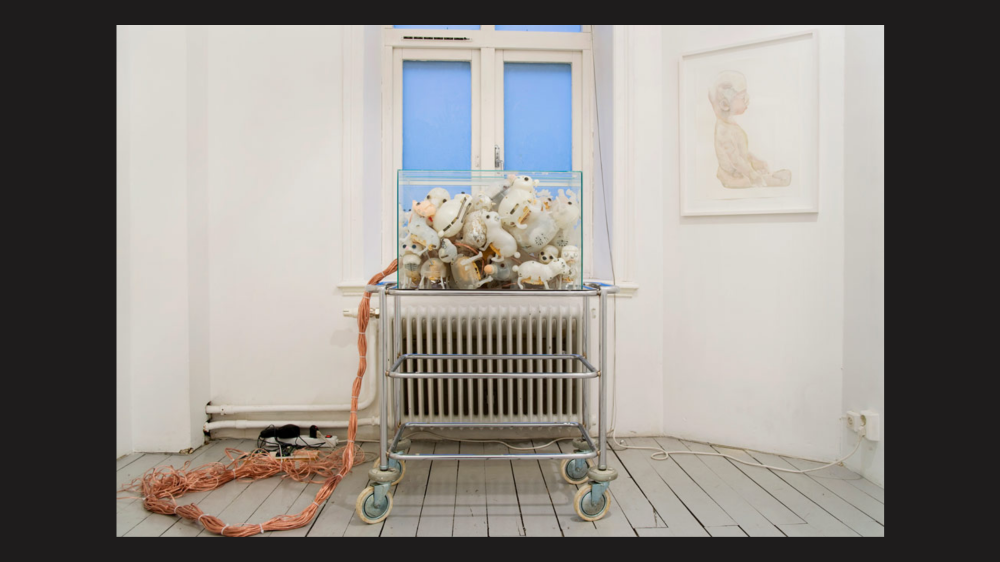 A glass case on a metal trolley. The class case is filled with toy animals whose fur has been removed. A thick line of cords is connected to the glass case, and there is a painting of a small child hanging on the white wall next to the glass case. There is a window directly behind the glass case.