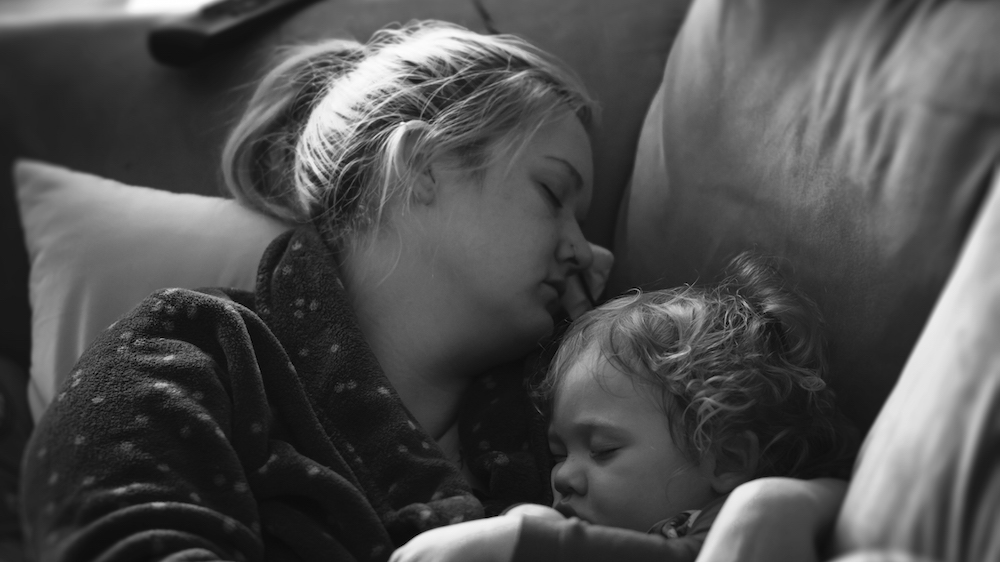 En mor liggerog sover på en sofa med et sykt barn